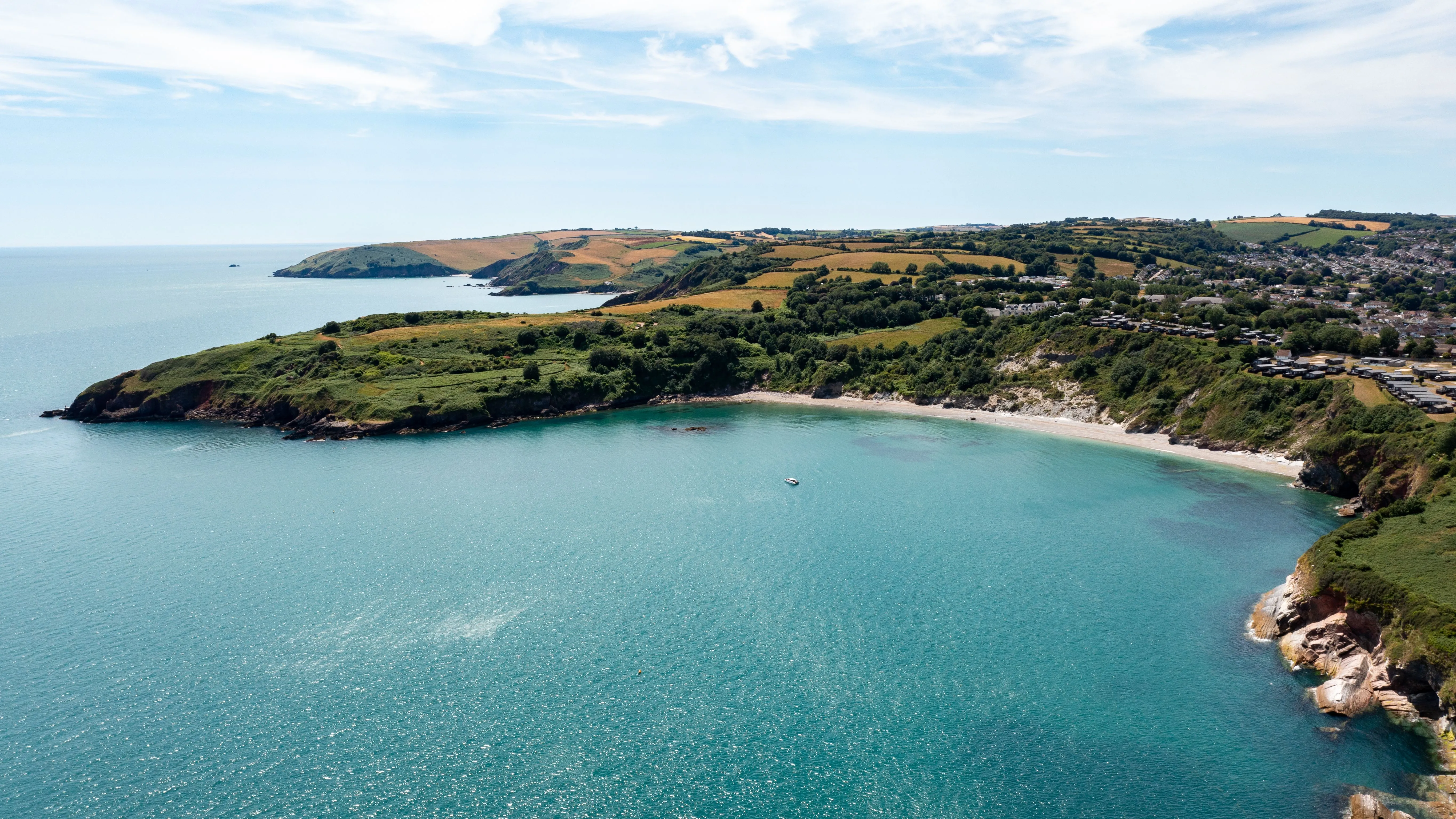 Aerial view of a turquoise coastal bay bordered by rocky cliffs, green headlands and patchwork farmland.