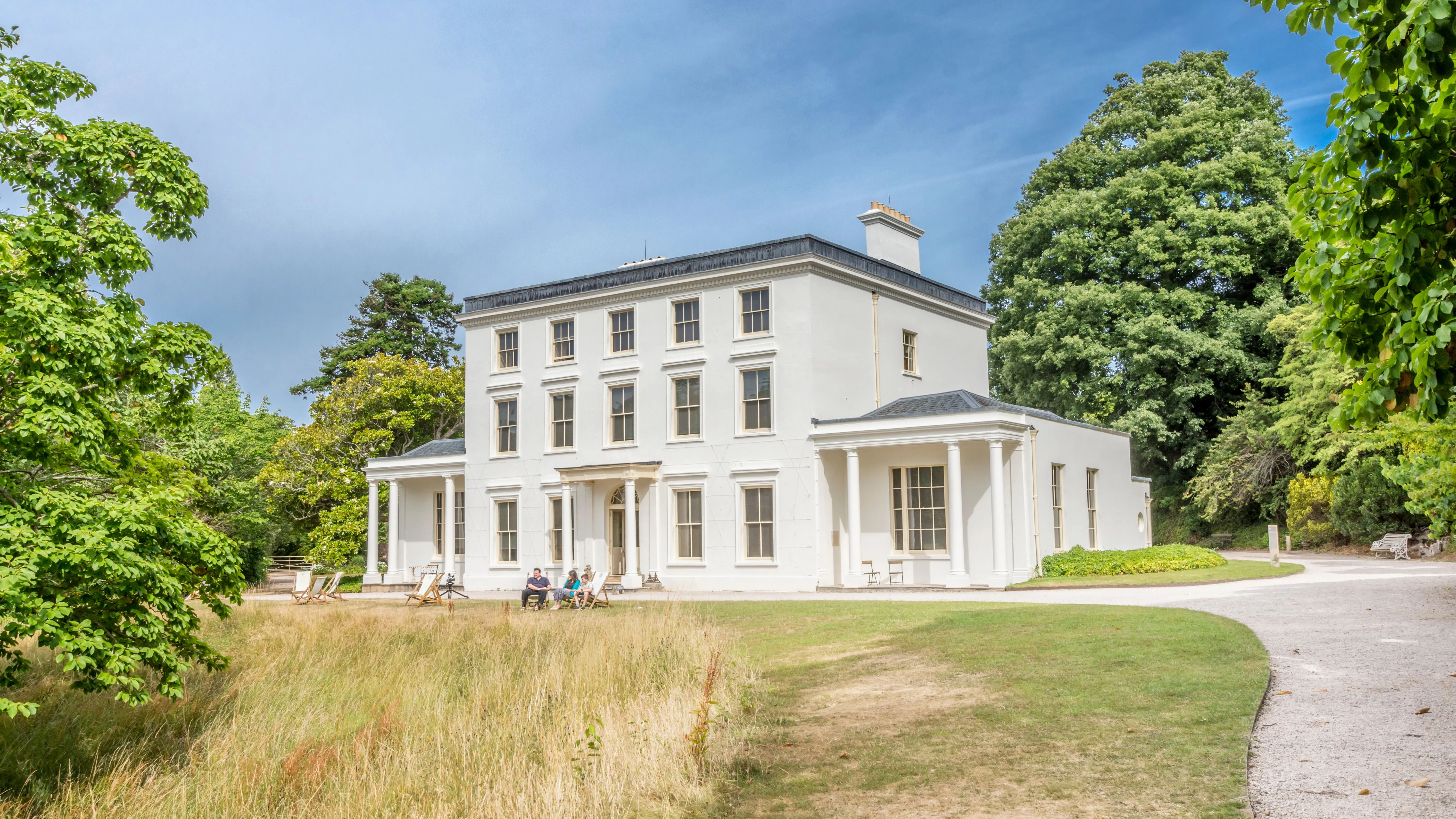 Greenway House, a white neoclassical country house surrounded by trees and lawns under a blue sky.