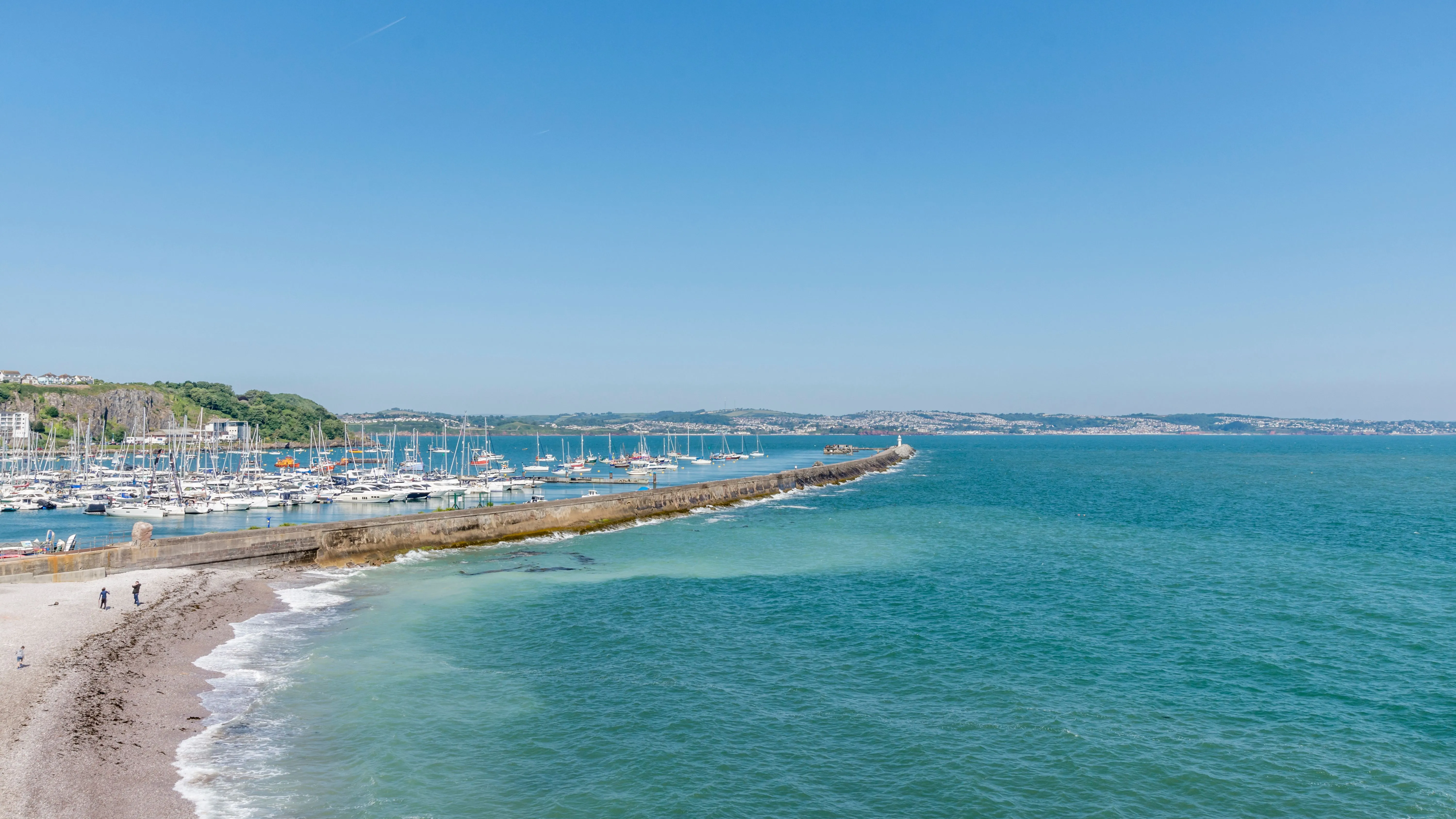 A long stone breakwater extends into turquoise sea beside a pebble beach and yacht marina under a clear blue sky.