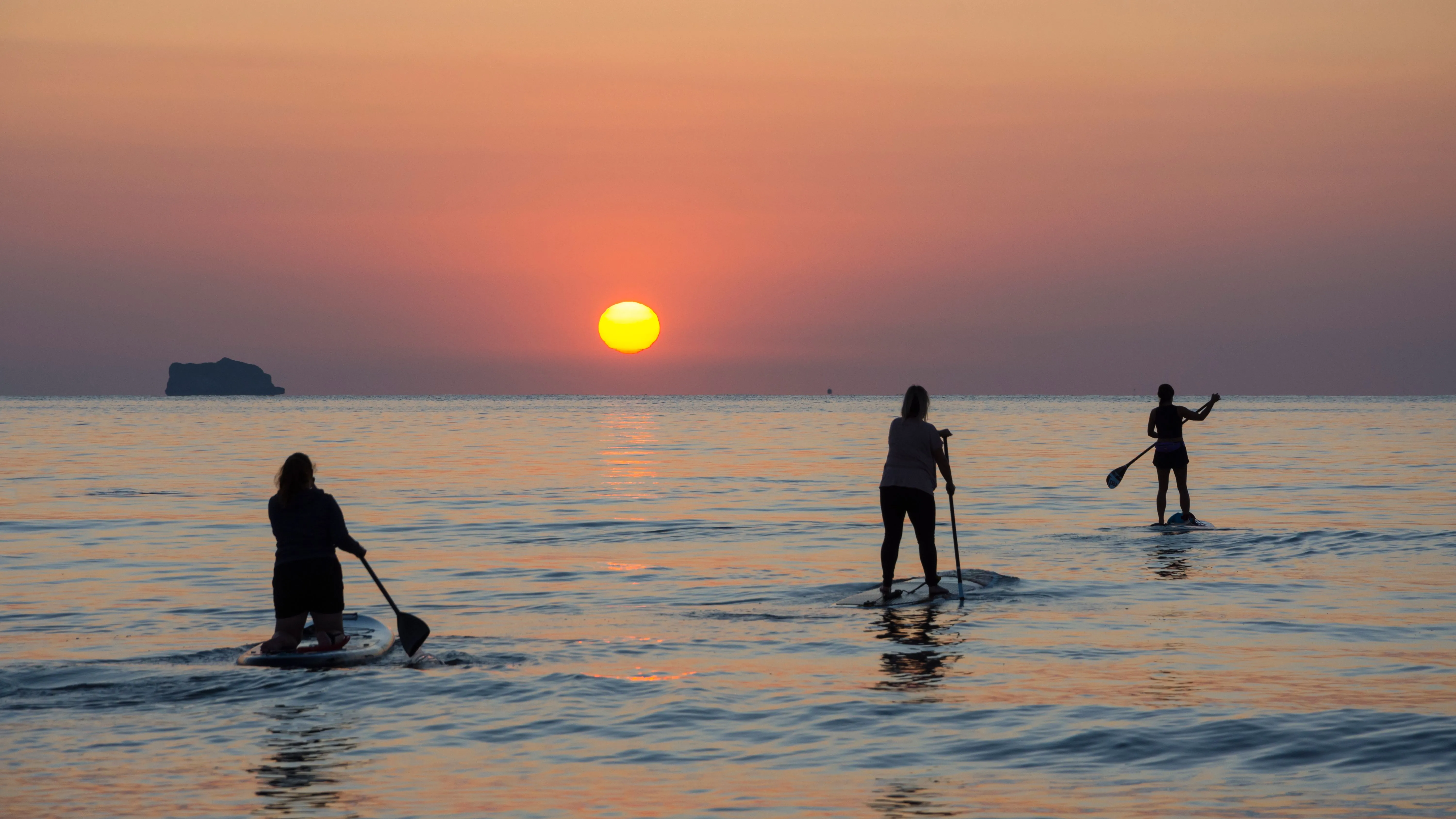 Three silhouetted people paddleboarding on calm sea water at sunrise, with a small island on the horizon.