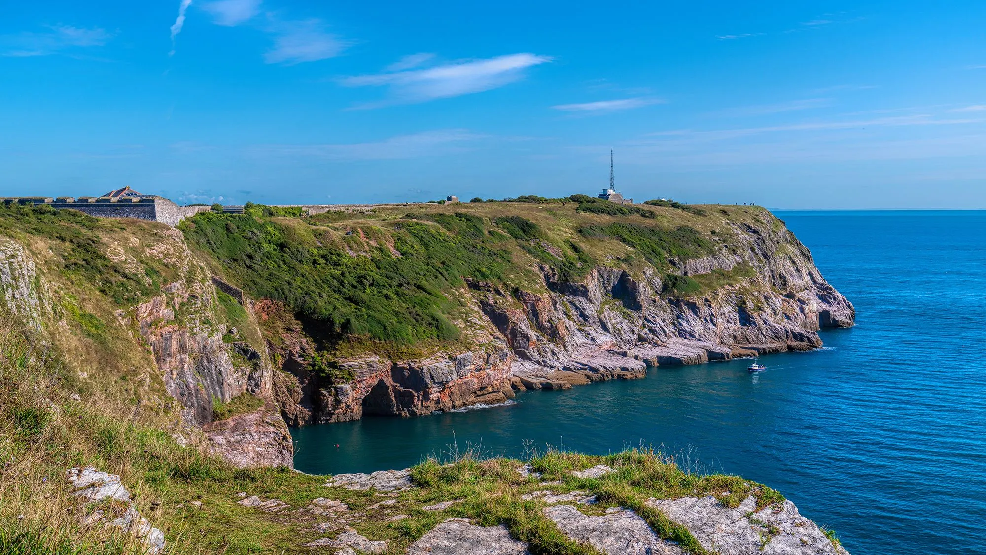 Rocky coastal cliffs with a historic fort and radio mast overlooking calm blue sea beneath a clear sky.