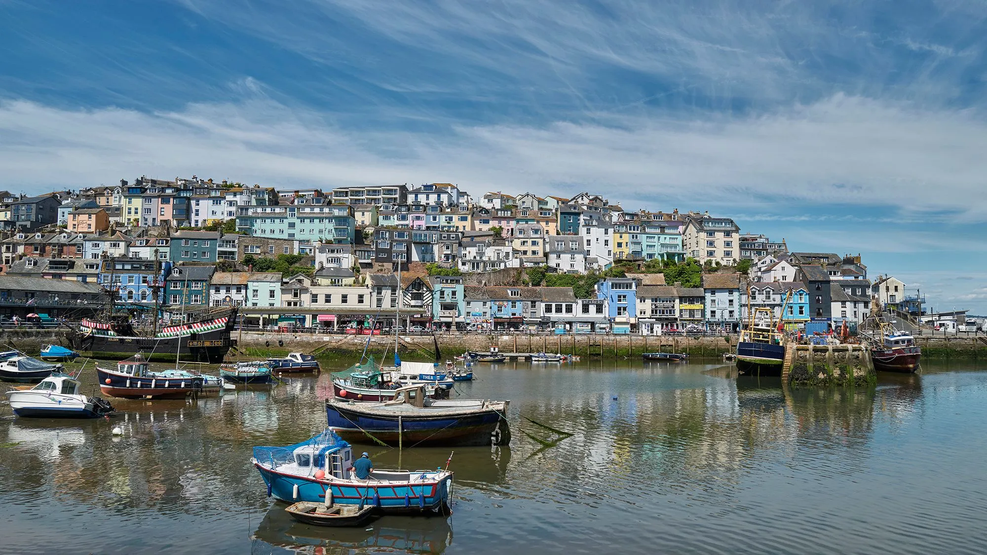 Fishing boats moored in Brixham harbour beneath colourful hillside houses and a blue sky.