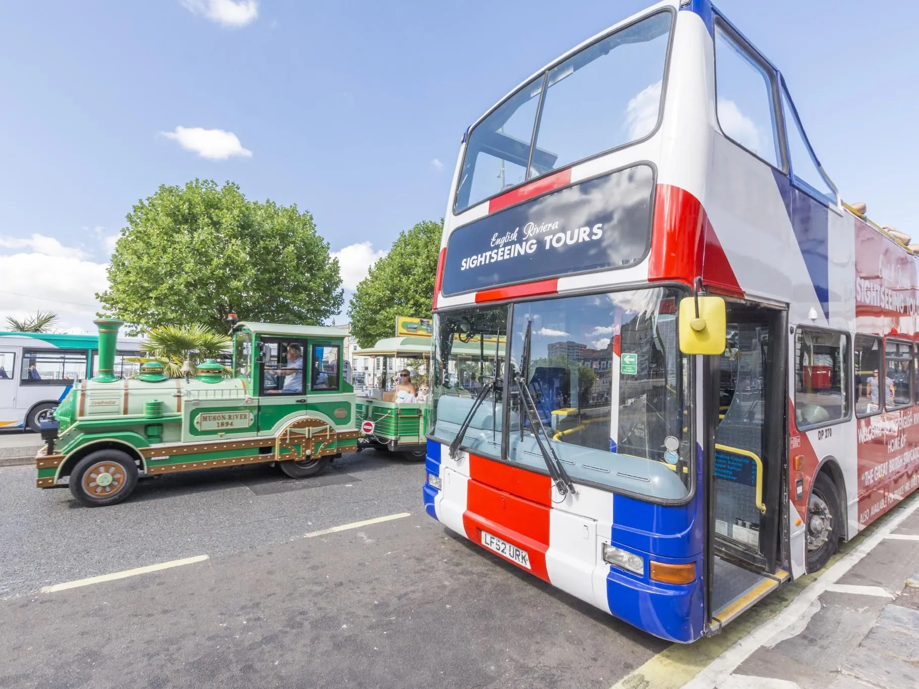 Open-top English Riviera sightseeing tour bus parked beside a green tourist road train on a sunny day