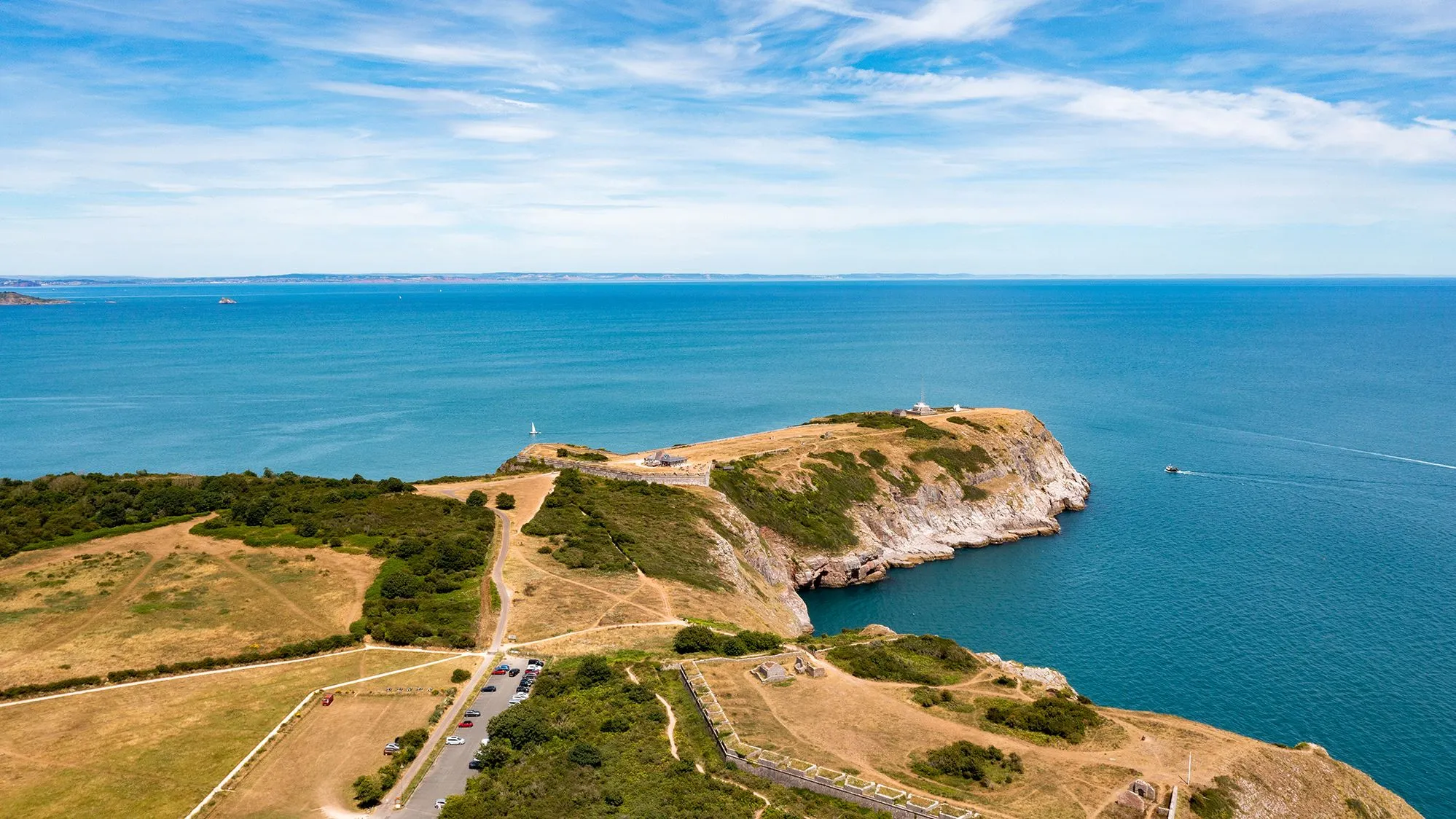 Aerial view of a rocky coastal headland with a lighthouse, footpaths and turquoise sea beneath a blue sky.
