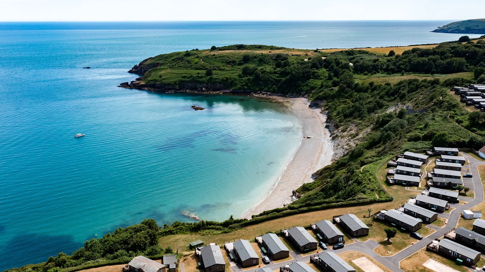 Aerial view of a sheltered sandy cove with turquoise sea, green cliffs and a coastal holiday park.