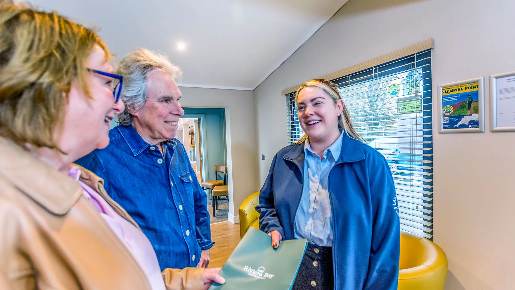 Smiling staff member welcomes an older couple in a bright reception area while handing over a River Bay folder.