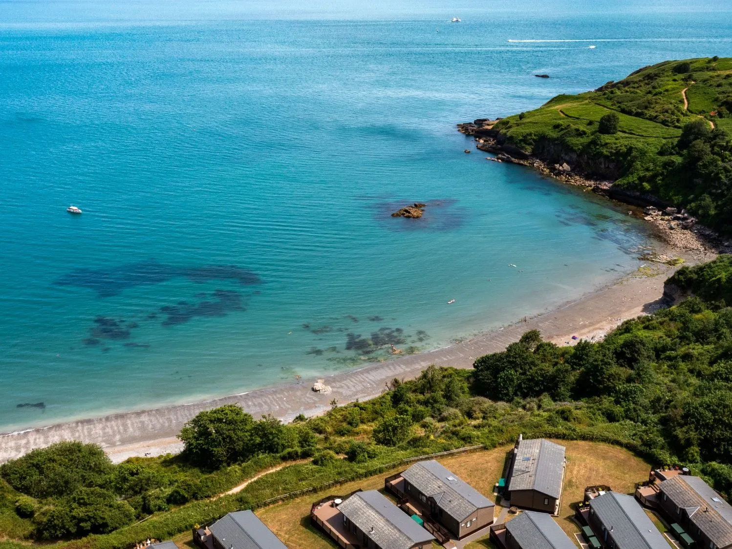 Aerial view of a turquoise coastal bay with a sandy beach, boats and green cliffs.