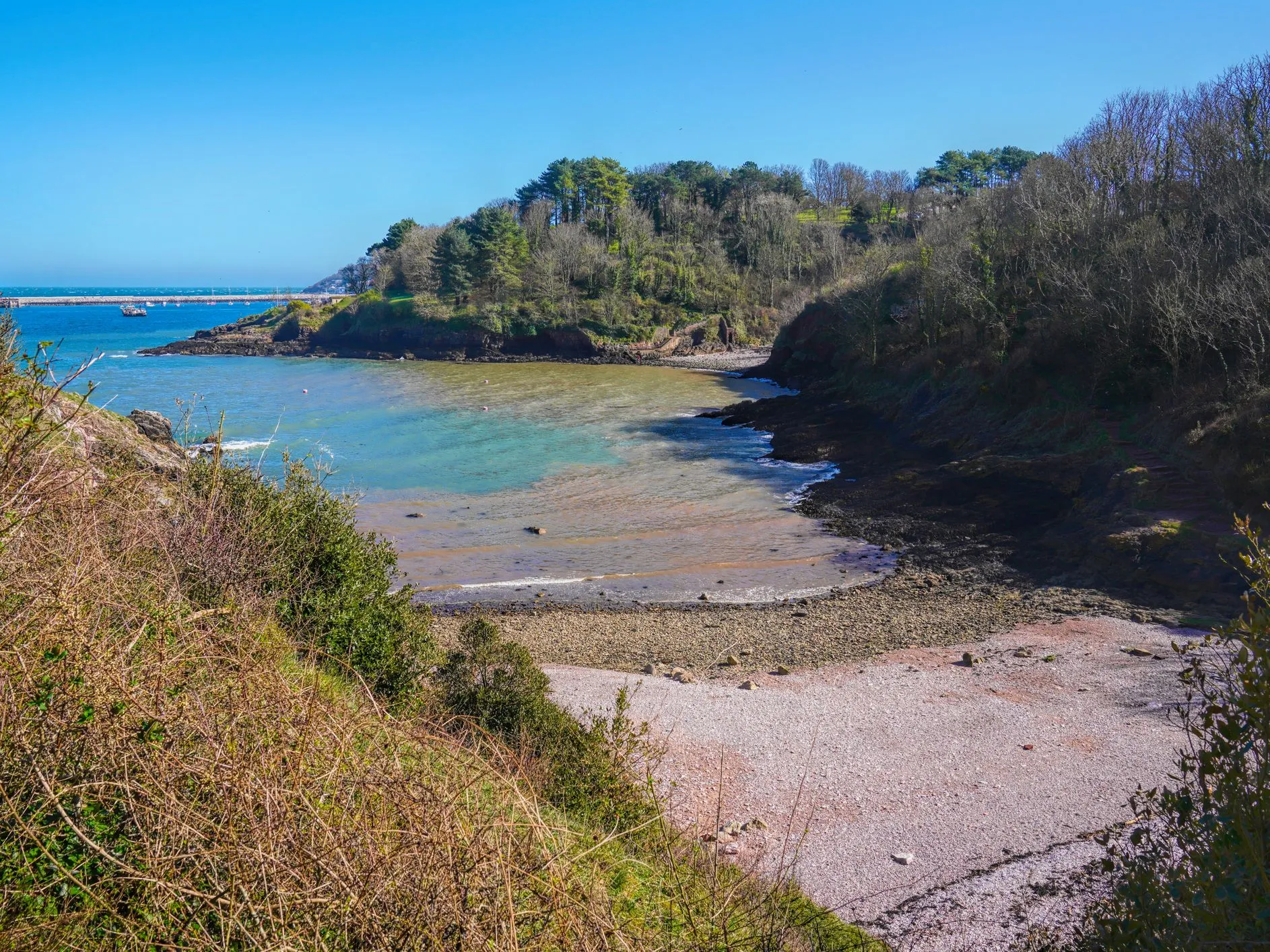 Sheltered pebble cove with clear turquoise water beneath wooded cliffs on a sunny day