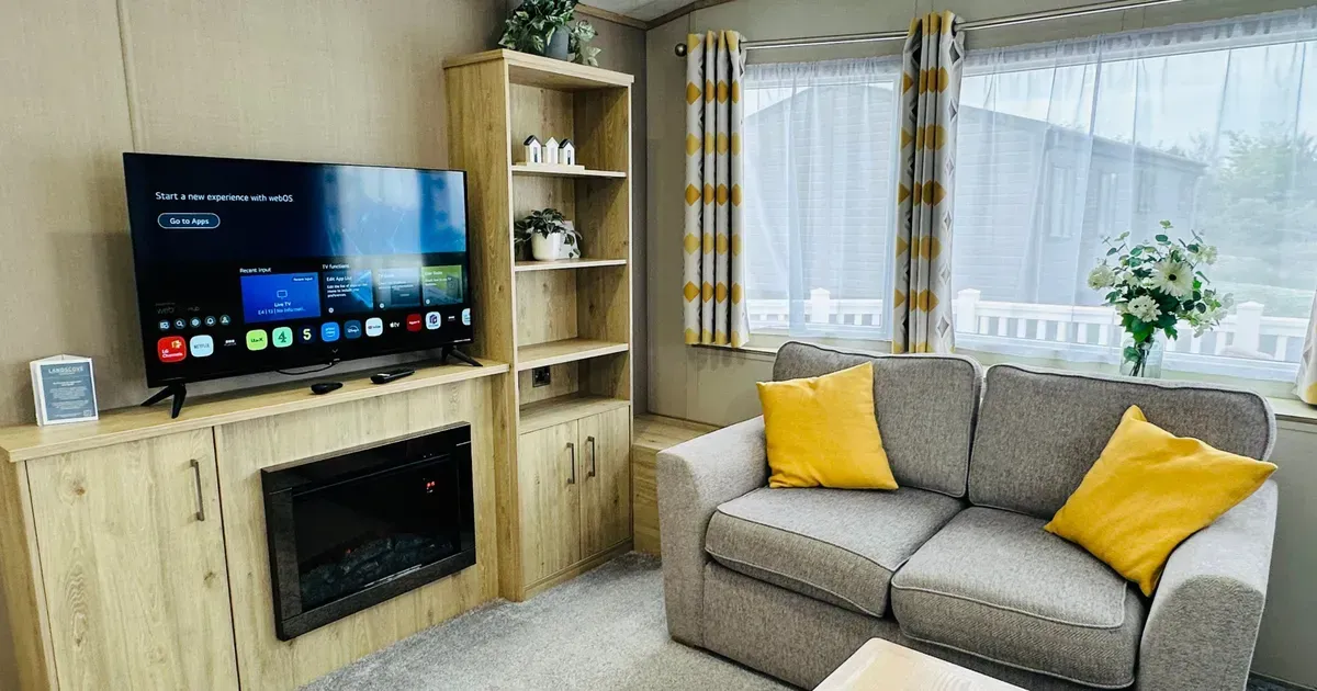 Cosy caravan lounge with grey sofa, yellow cushions, television and electric fireplace beside a large window.