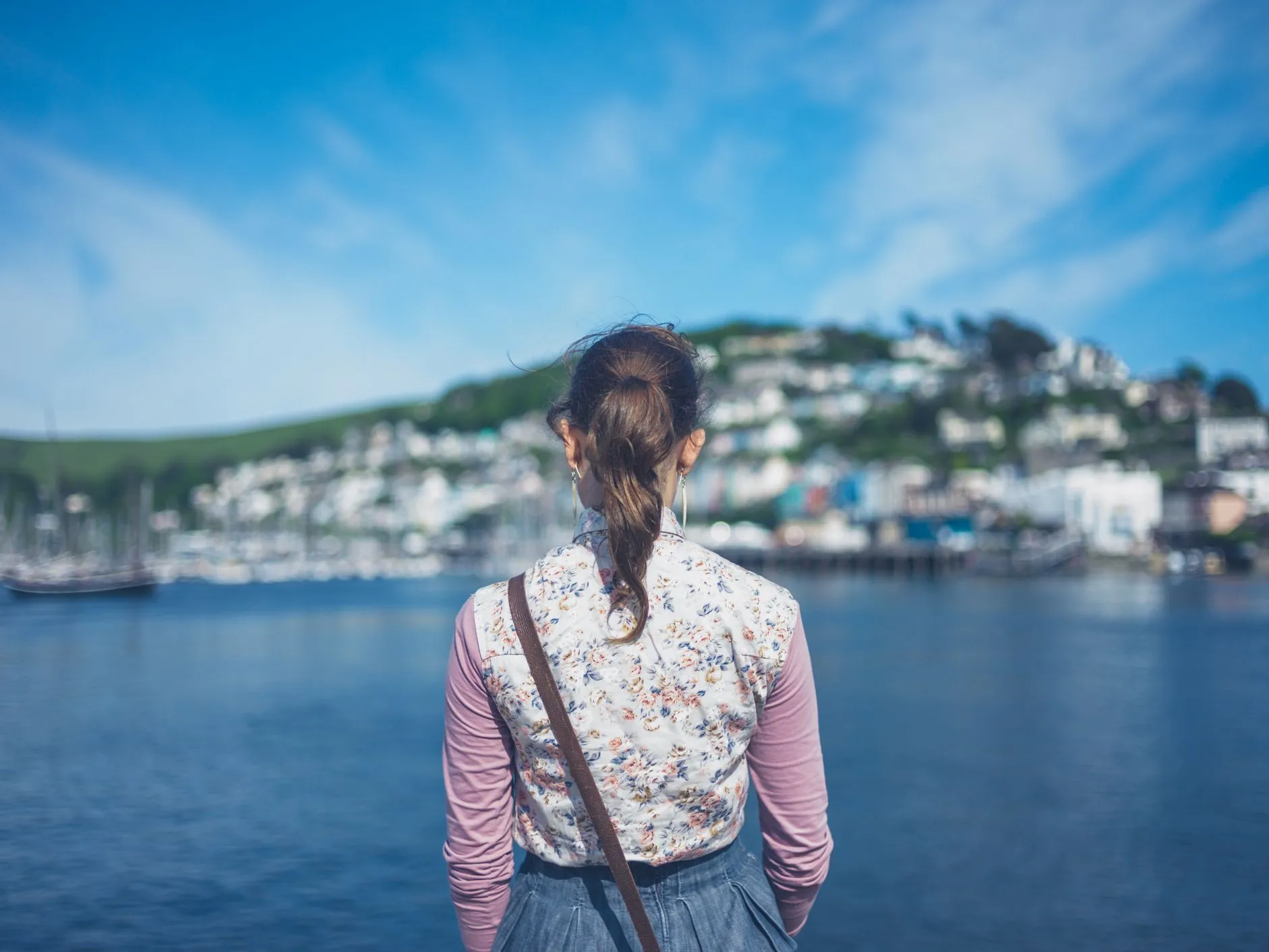 Woman viewed from behind looking across a harbour towards a coastal hillside town under a blue sky