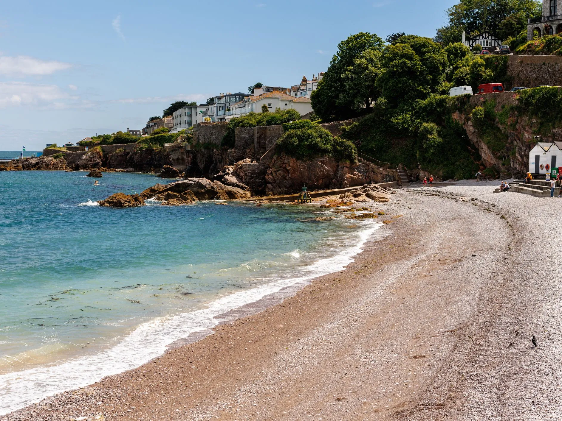 Pebble beach with turquoise sea beneath houses and greenery on a rocky coastal hillside