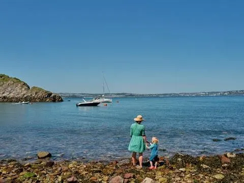 Adult and child on a rocky shoreline at Fishcombe Cove, looking out over calm blue sea with a moored sailboat