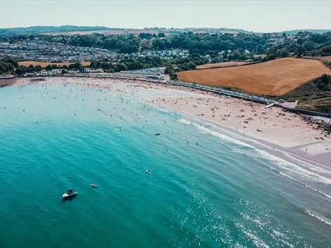 Aerial view of a sandy beach with swimmers, boats on turquoise sea, and rolling countryside beyond