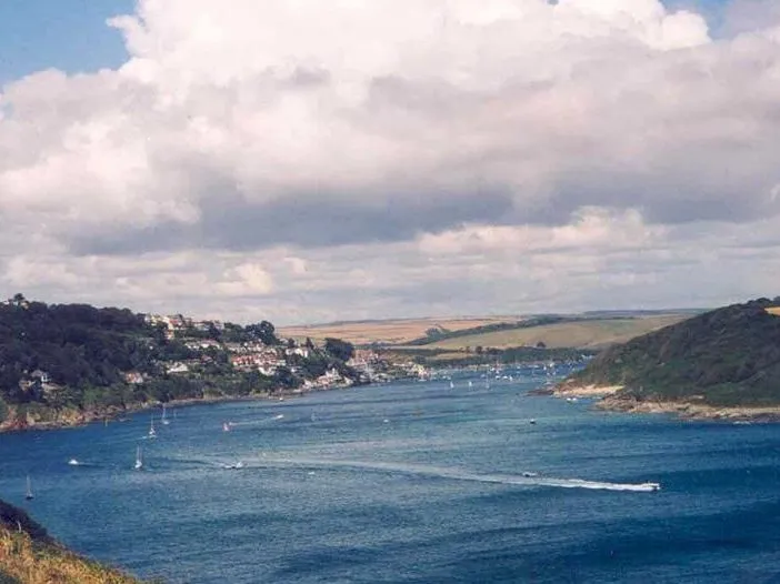 Wide view of a coastal estuary with sailboats and a speedboat wake, wooded hills and a village under a cloudy sky