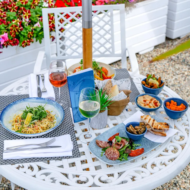 Outdoor dining table on the Ommaroo food terrace with pasta, charcuterie, bread, dips and glasses of wine beside bright flower planters.
