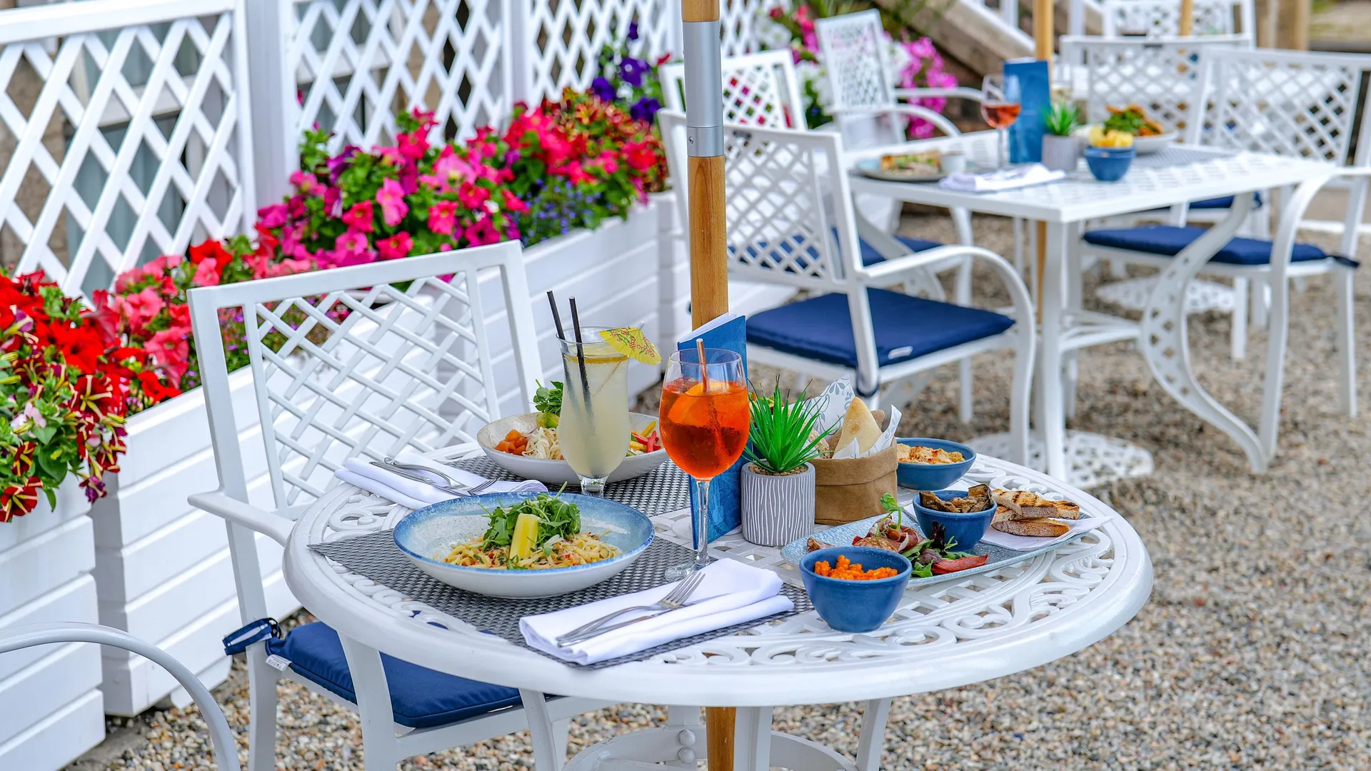 Outdoor food terrace with white tables and chairs, colourful flower planters, and a table set with drinks and dishes.