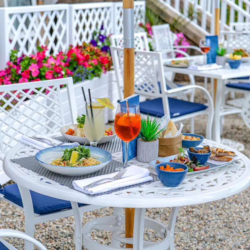 Outdoor food terrace with white tables and chairs, colourful flowers, plated dishes and cocktails