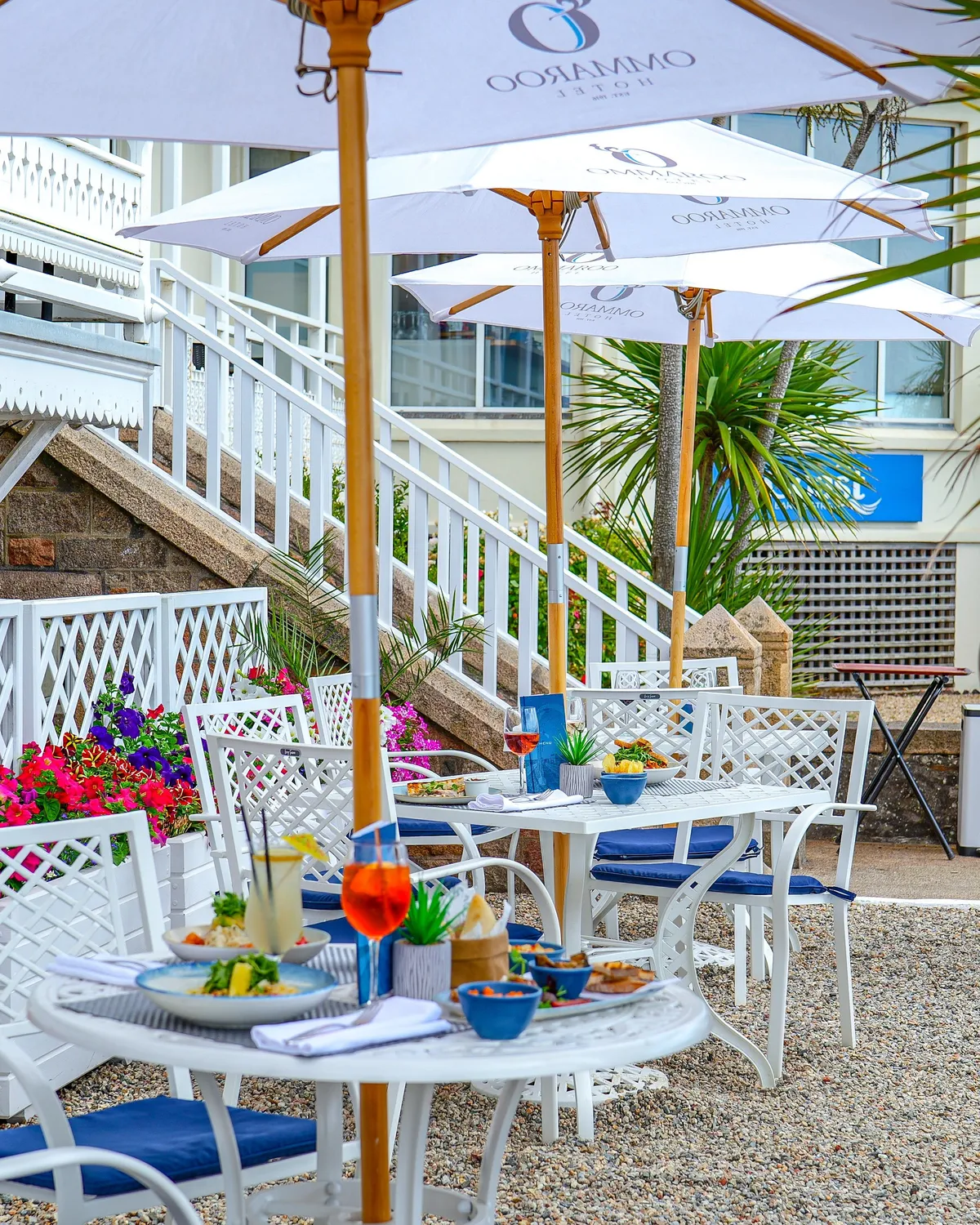 Outdoor dining terrace at Ommaroo Hotel with white umbrellas, tables set with food and drinks, and bright flower planters