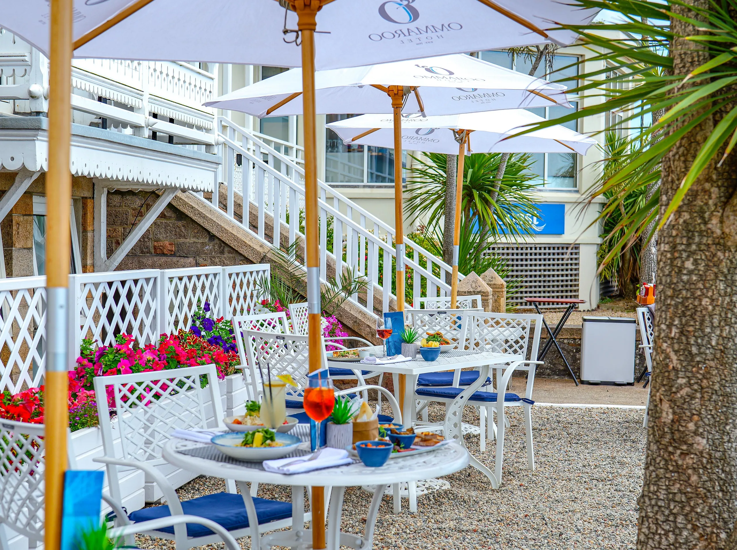 Outdoor dining terrace at Ommaroo Hotel with white umbrellas, tables set with food and drinks, and bright flower planters
