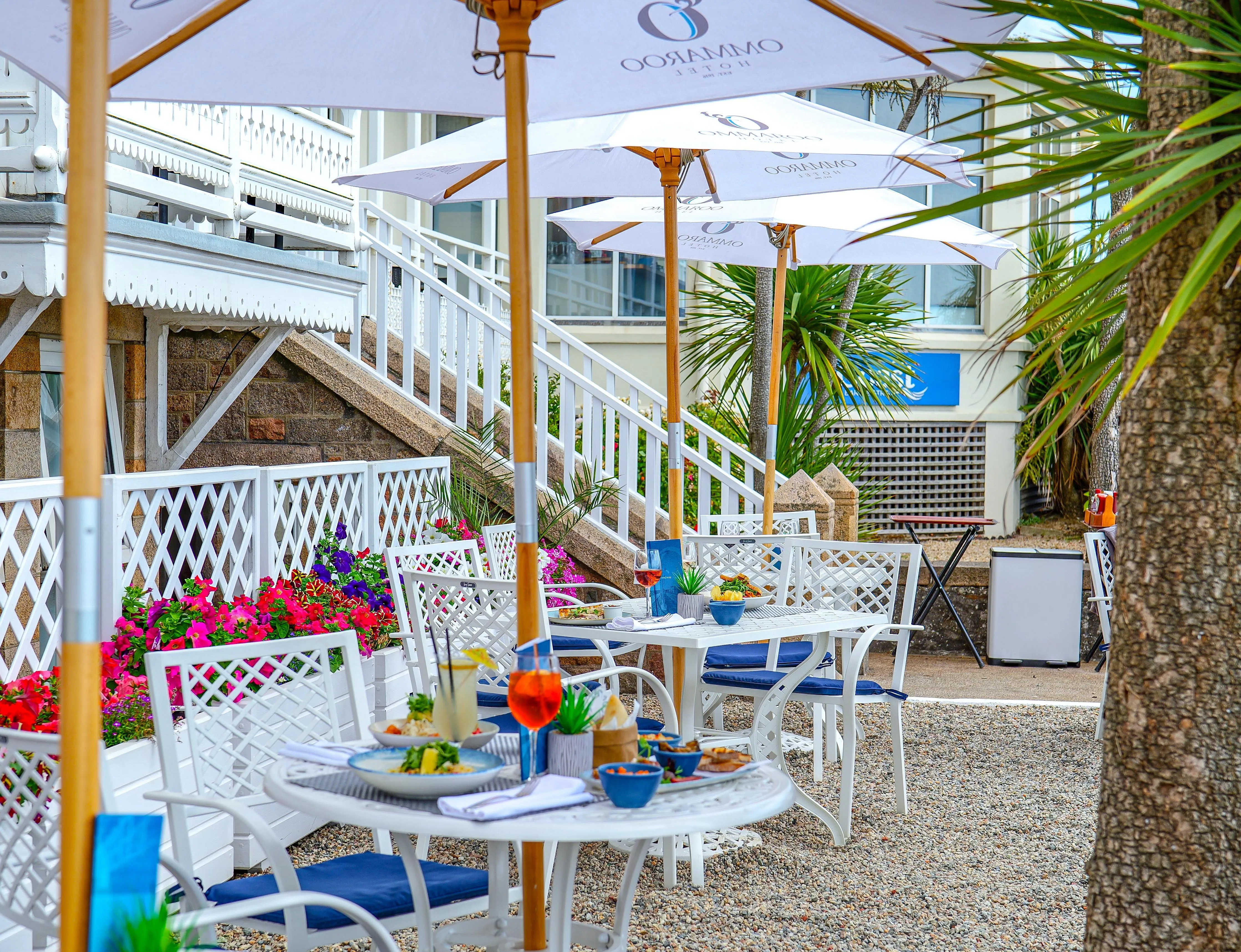 Outdoor dining terrace at Ommaroo Hotel with white umbrellas, tables set with food and drinks, and bright flower planters