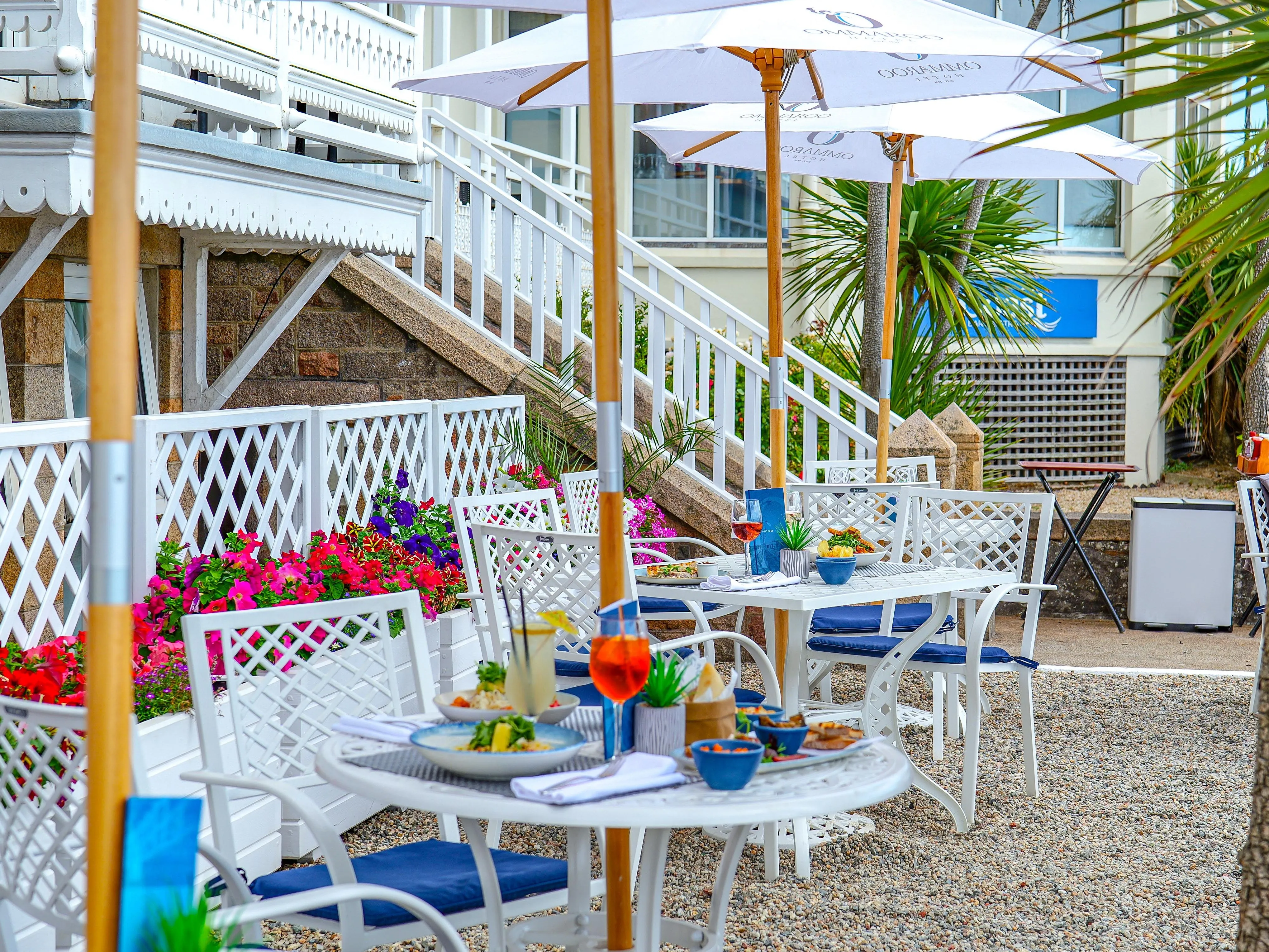 Outdoor dining terrace at Ommaroo Hotel with white umbrellas, tables set with food and drinks, and bright flower planters