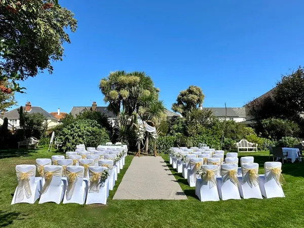 Outdoor wedding ceremony setup in a garden with white chairs, gold sashes and a floral arch under a clear blue sky