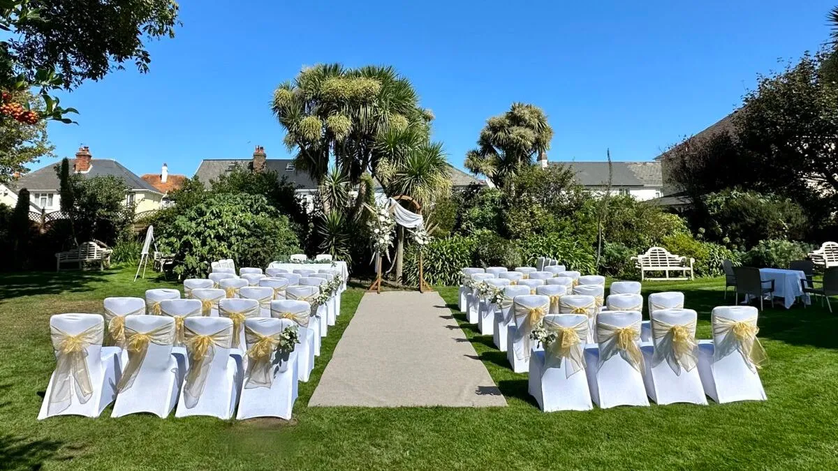 Outdoor wedding ceremony setup in a garden with white chairs, gold sashes and a floral arch under a clear blue sky