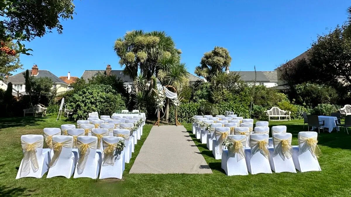 Outdoor wedding ceremony setup in a garden with white chairs, gold sashes and a floral arch under a clear blue sky