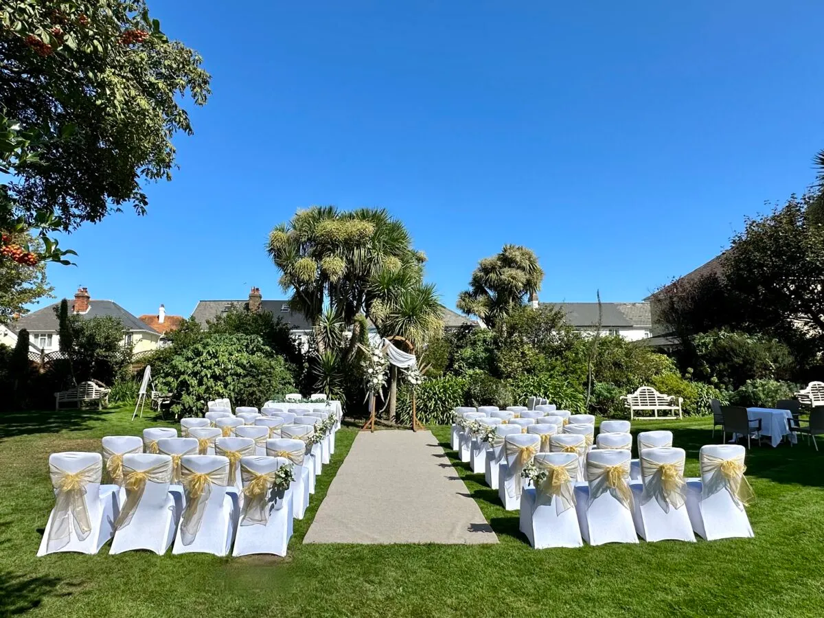Outdoor wedding ceremony setup in a garden with white chairs, gold sashes and a floral arch under a clear blue sky