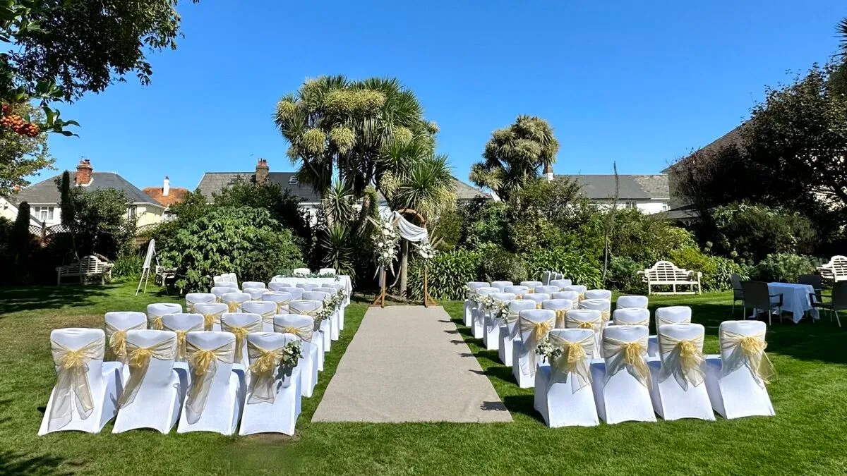 Outdoor wedding ceremony setup in a garden with white chairs, gold sashes and a floral arch under a clear blue sky