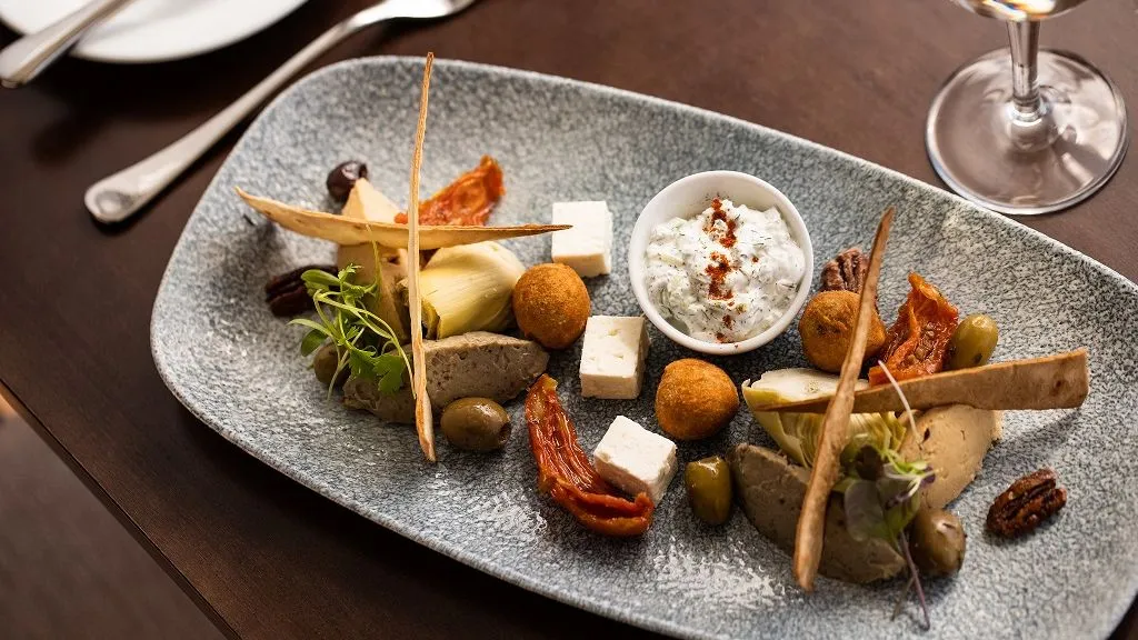 Mediterranean-style starter platter with olives, feta, artichokes, sun-dried tomatoes, crisp flatbread and a creamy dip on a speckled serving plate