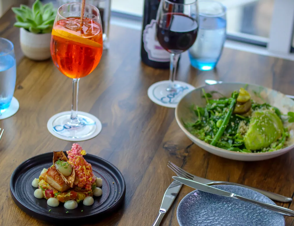 Elegant restaurant table with a plated main course, green salad, an orange spritz and a glass of red wine by the window