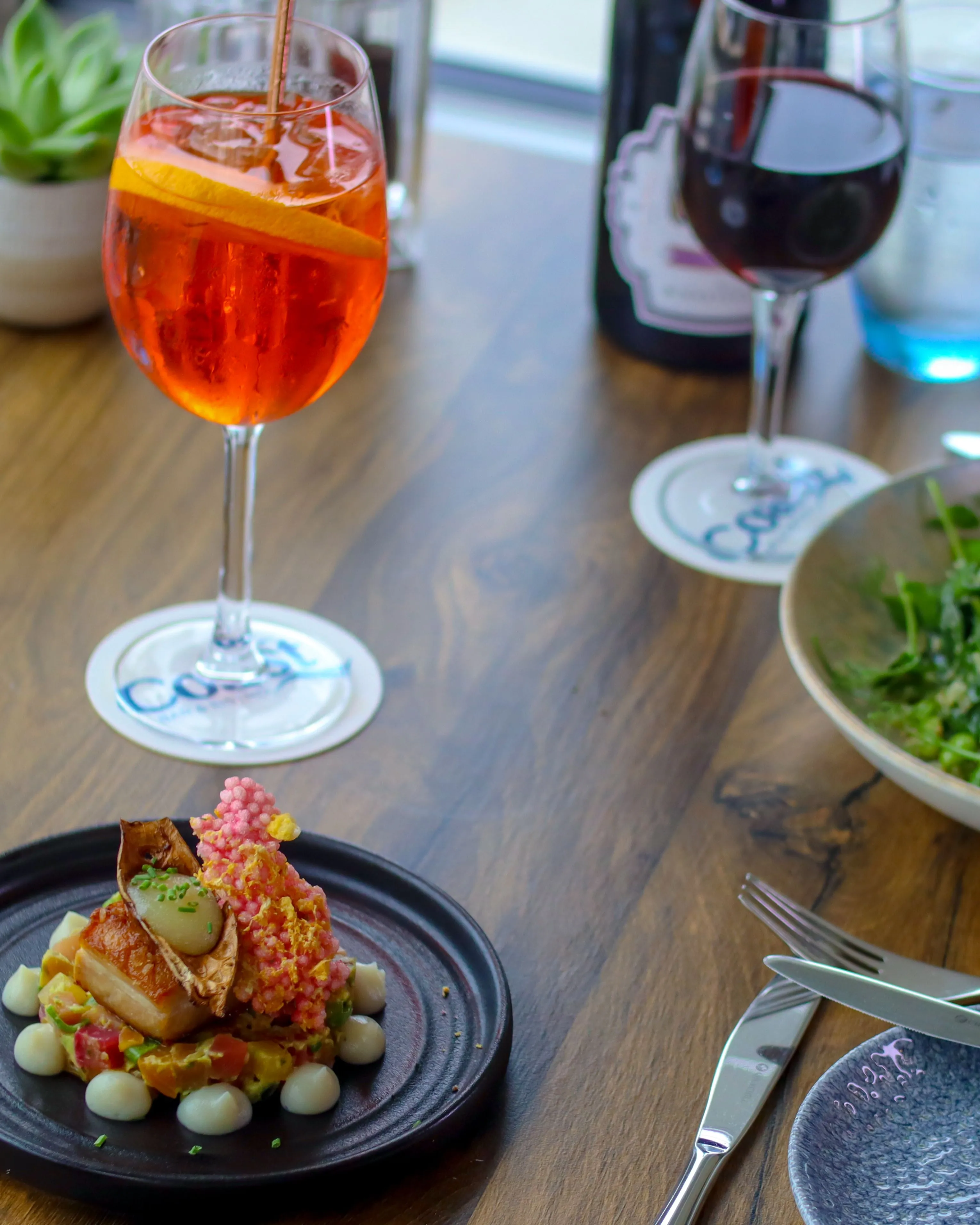 Elegant restaurant table with a plated main course, green salad, an orange spritz and a glass of red wine by the window
