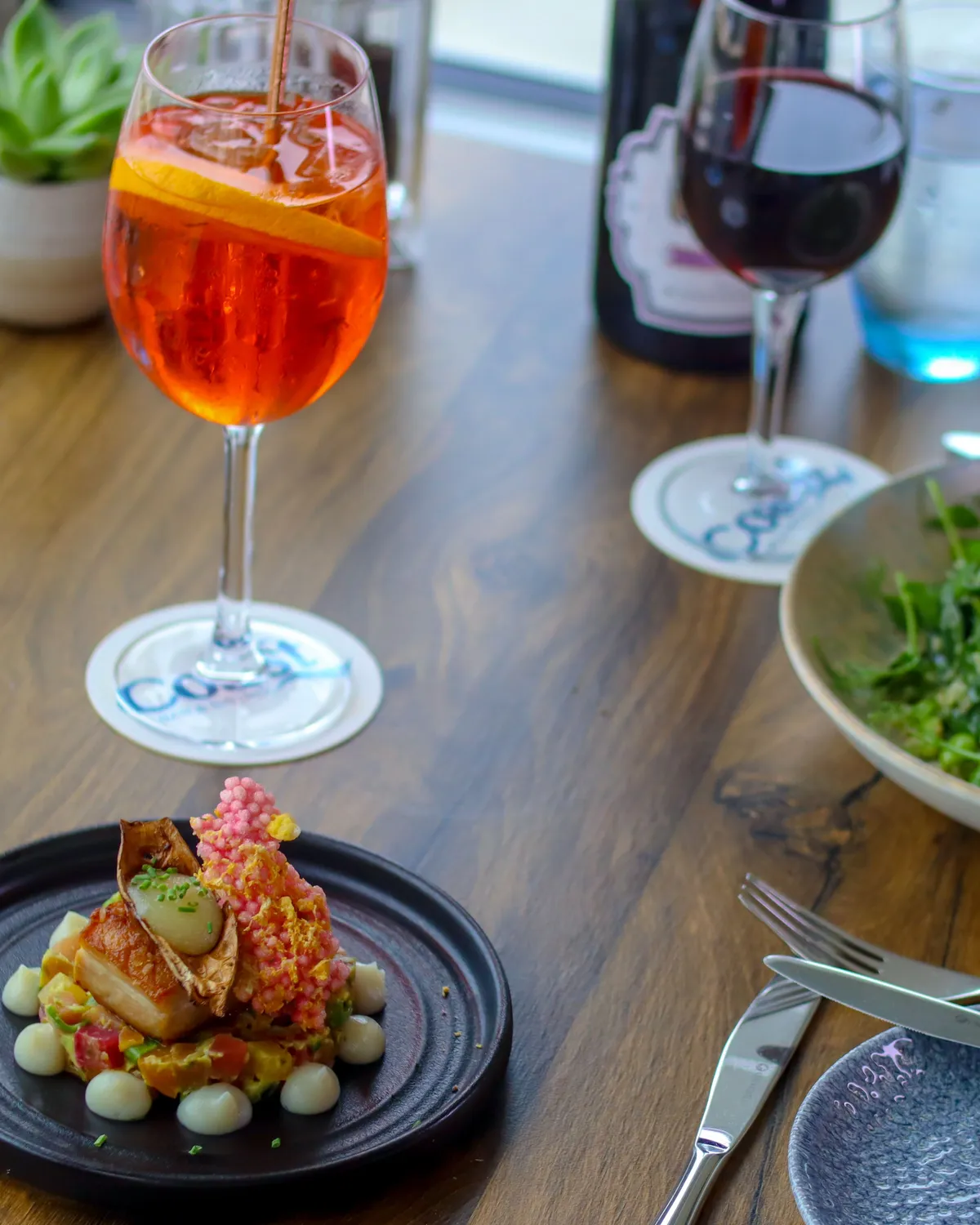 Elegant restaurant table with a plated main course, green salad, an orange spritz and a glass of red wine by the window