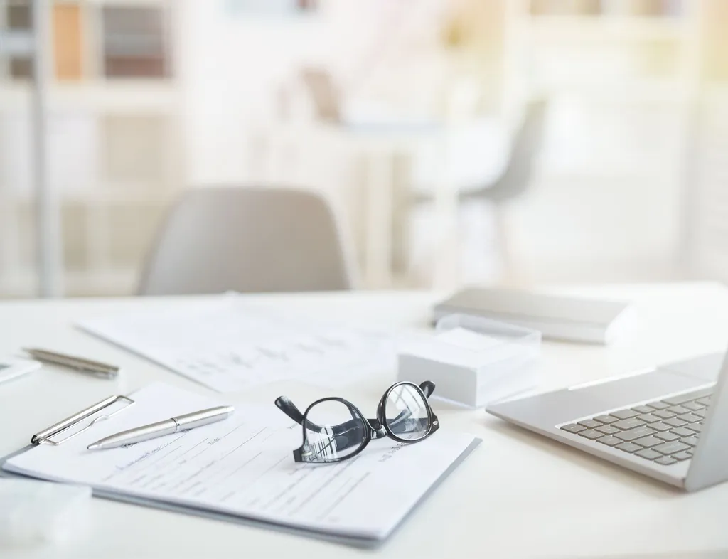Bright office desk with paperwork, spectacles, pen, tablet and laptop