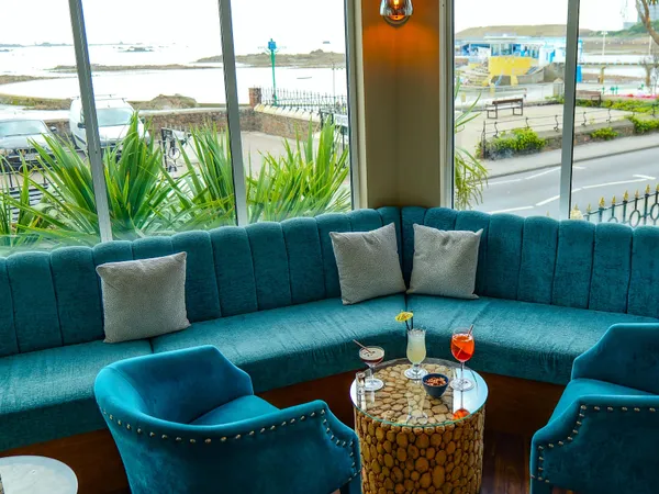 Stylish bar seating area with teal sofas and chairs beside large windows overlooking the seafront.