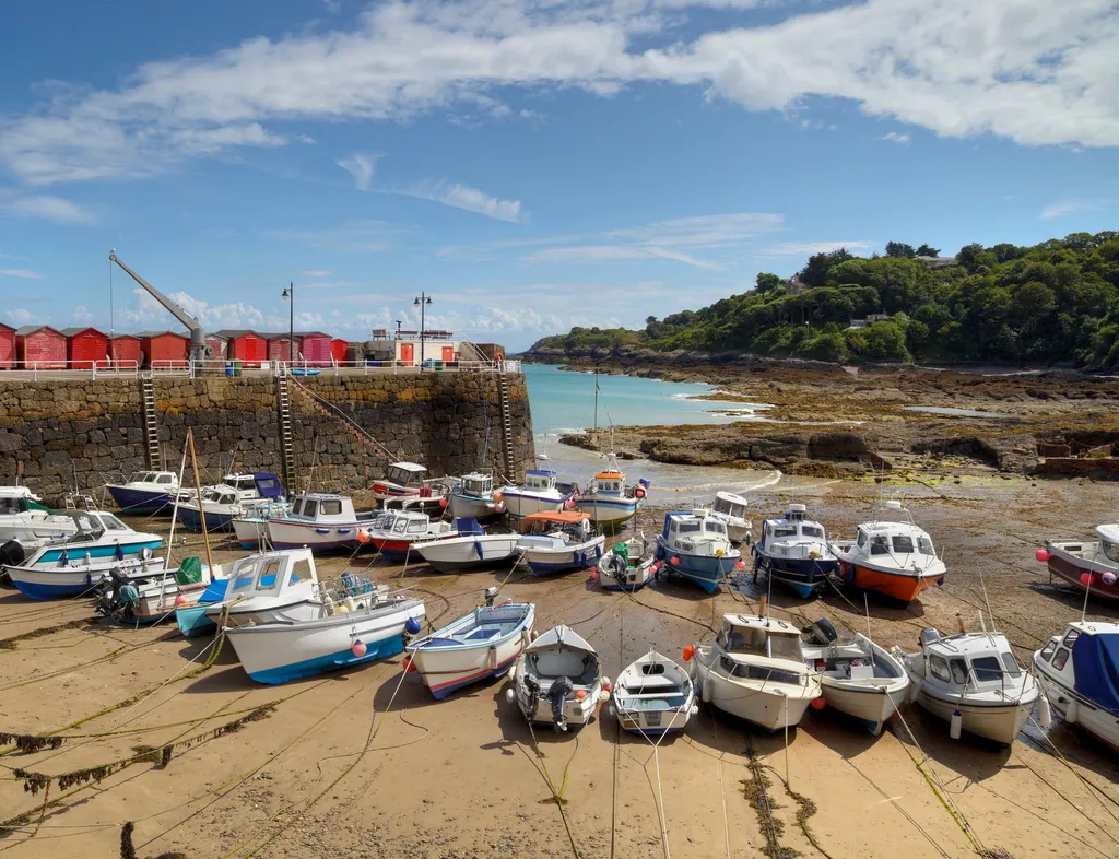 Small fishing boats resting on the sand at low tide in Rozel Bay, with red harbour sheds, a stone quay and a wooded headland under a bright blue sky.