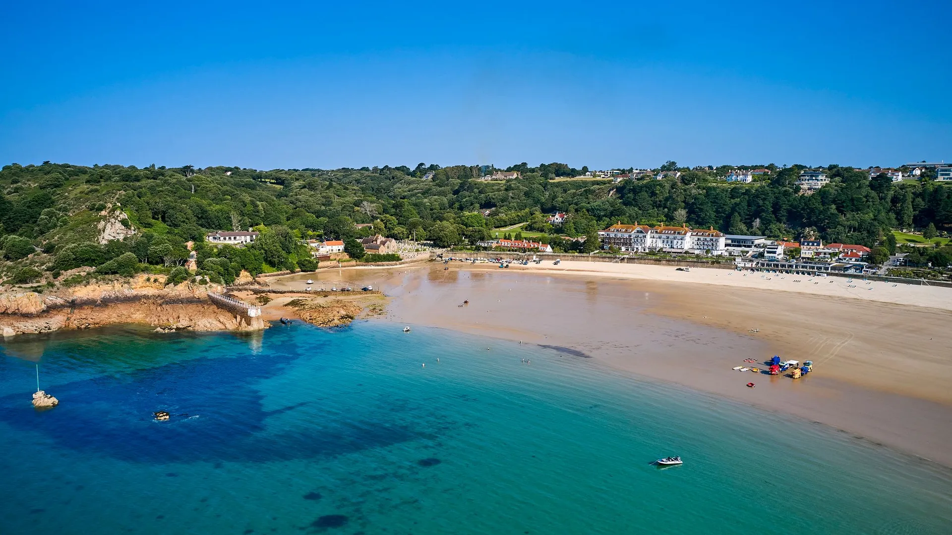 Aerial view of St Brelades Bay with turquoise water, sandy beach and coastal buildings beneath a clear blue sky.