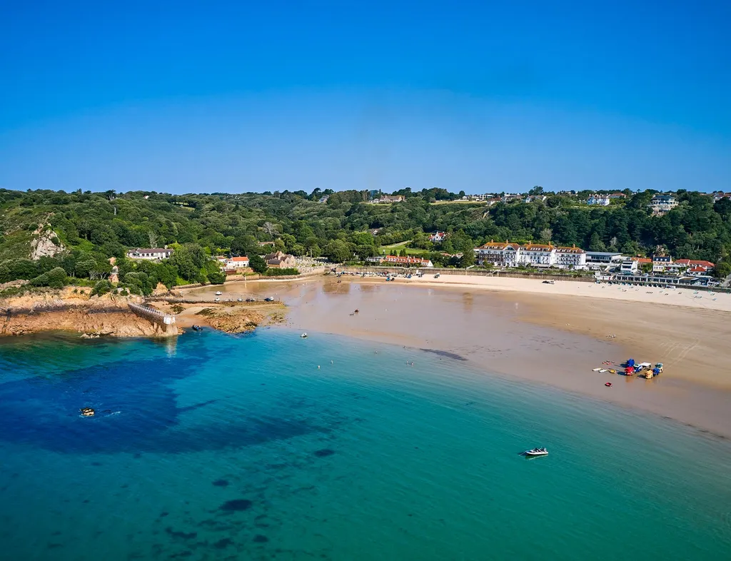 Aerial view of St Brelades Bay with turquoise water, sandy beach and coastal buildings beneath a clear blue sky.
