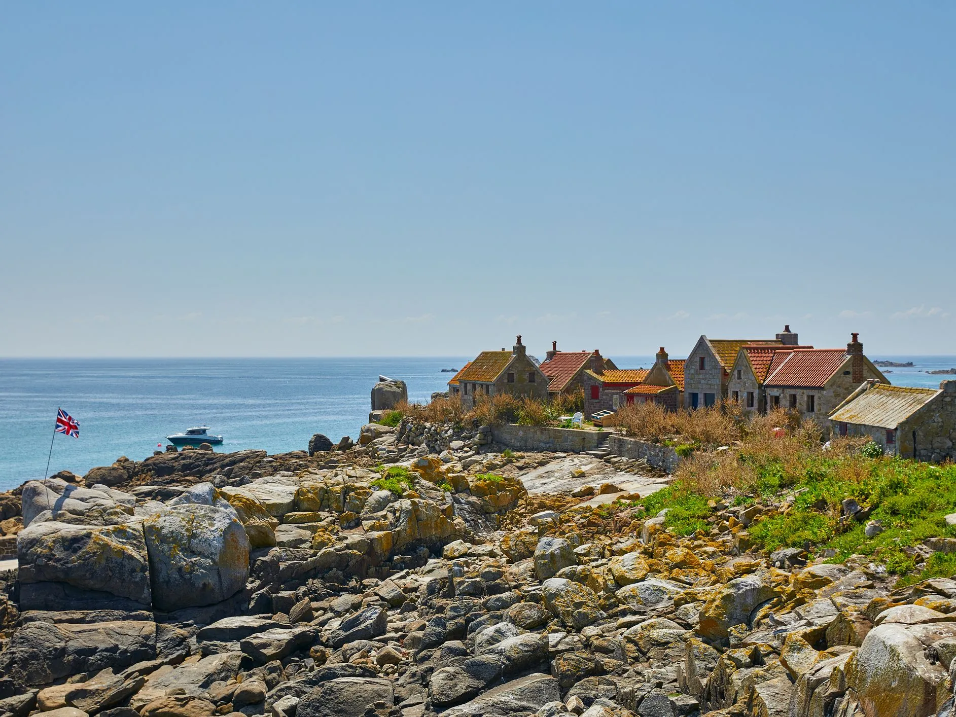 Stone cottages on a rocky island coastline overlooking a calm blue sea, with a Union Jack flag and a small boat offshore.