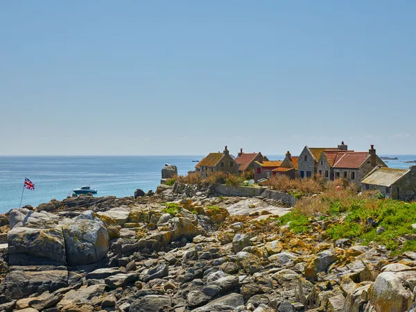 Stone cottages on a rocky island coastline overlooking a calm blue sea, with a Union Jack flag and a small boat offshore.