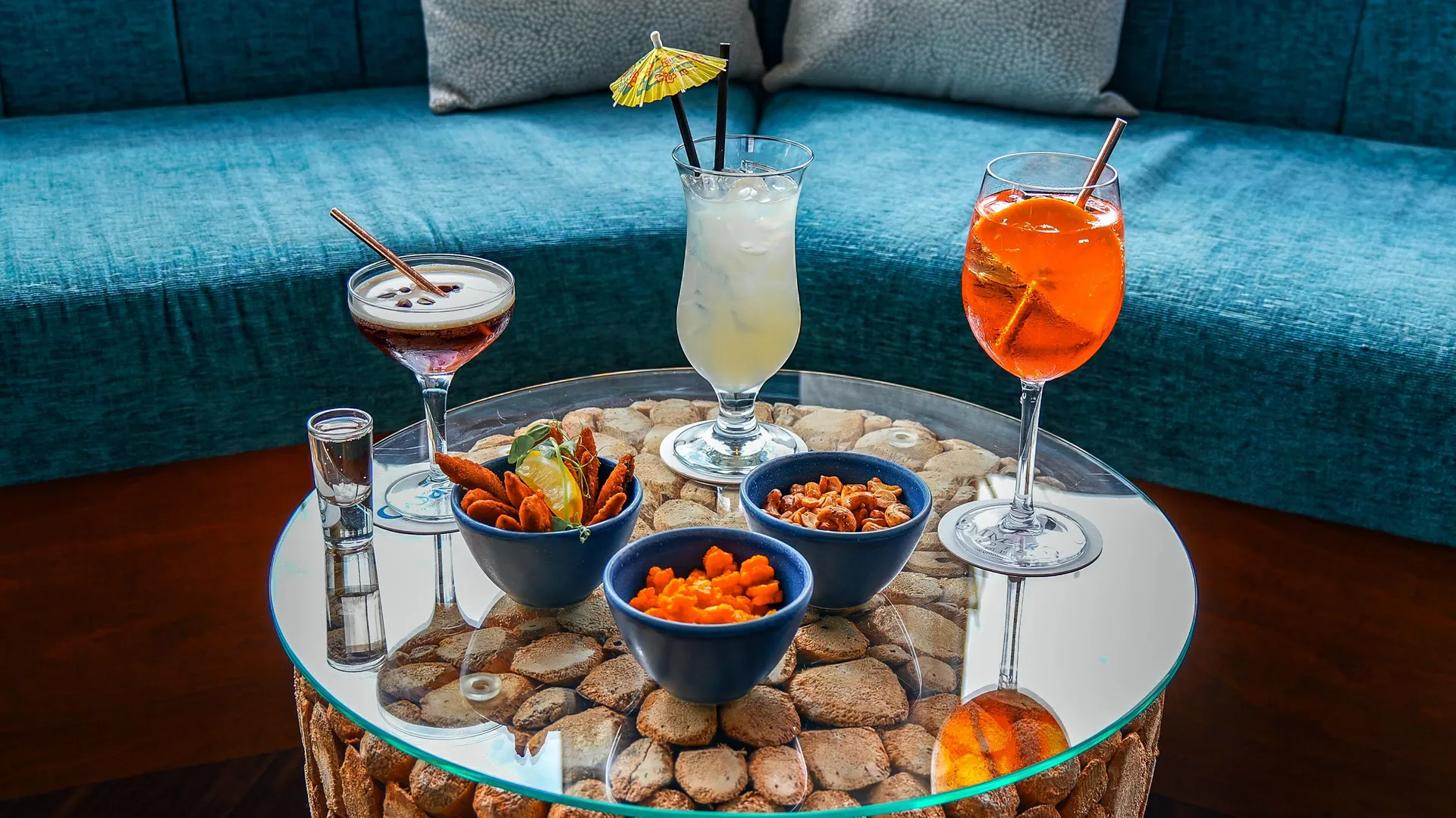 A selection of cocktails and bar snacks on a glass-topped table in a stylish lounge with blue seating.