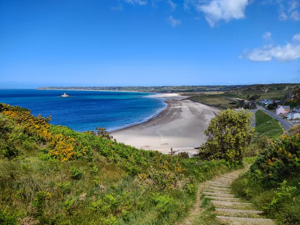 Panoramic view of St Ouen's Bay with sandy beach, turquoise sea and coastal path descending through green hillside
