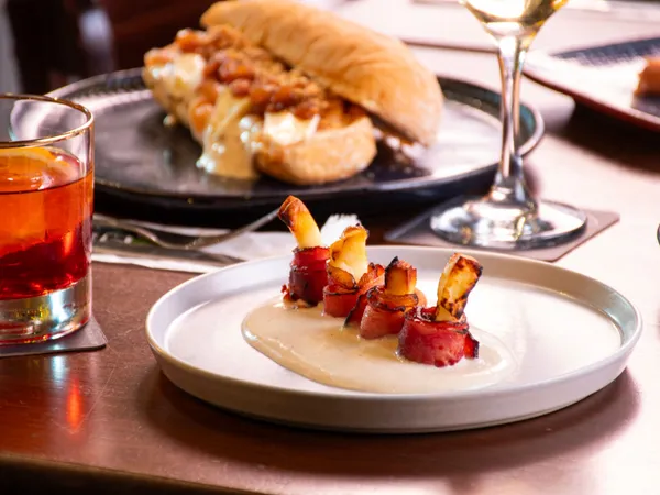 Plated bacon-wrapped appetisers on a creamy sauce with a hot dog and drinks on a restaurant table