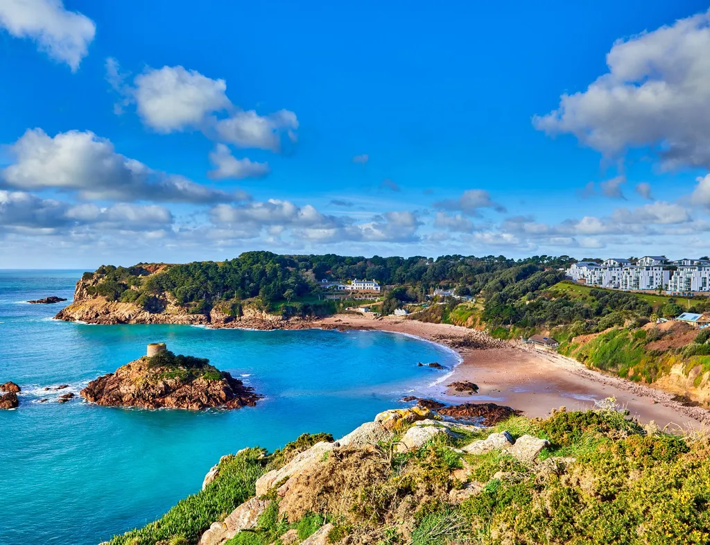 Coastal bay with turquoise water, sandy beach and a small rocky islet topped by a historic tower under a bright blue sky