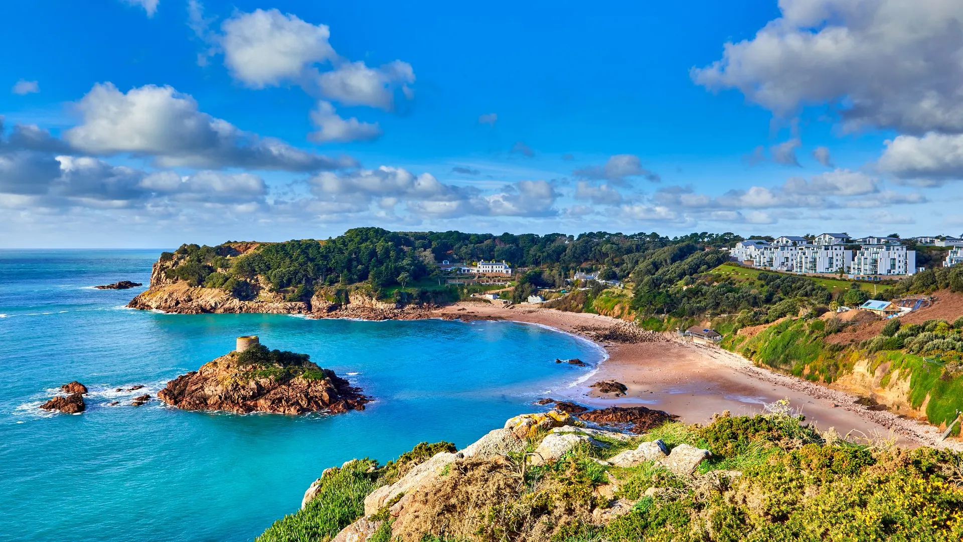 Coastal bay with turquoise water, sandy beach and a small rocky islet topped by a historic tower under a bright blue sky
