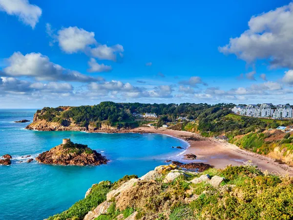 Coastal bay with turquoise water, sandy beach and a small rocky islet topped by a historic tower under a bright blue sky