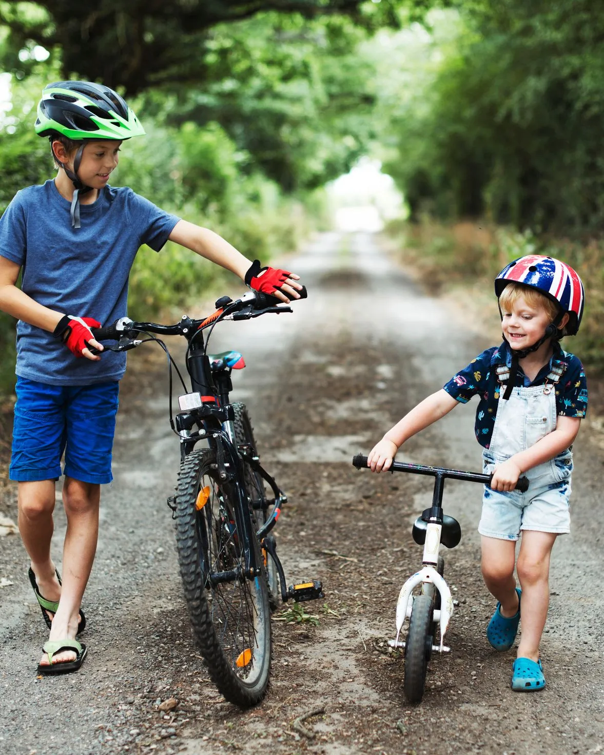 Two children wearing helmets walk with a bicycle and a balance bike along a quiet country lane lined with trees.