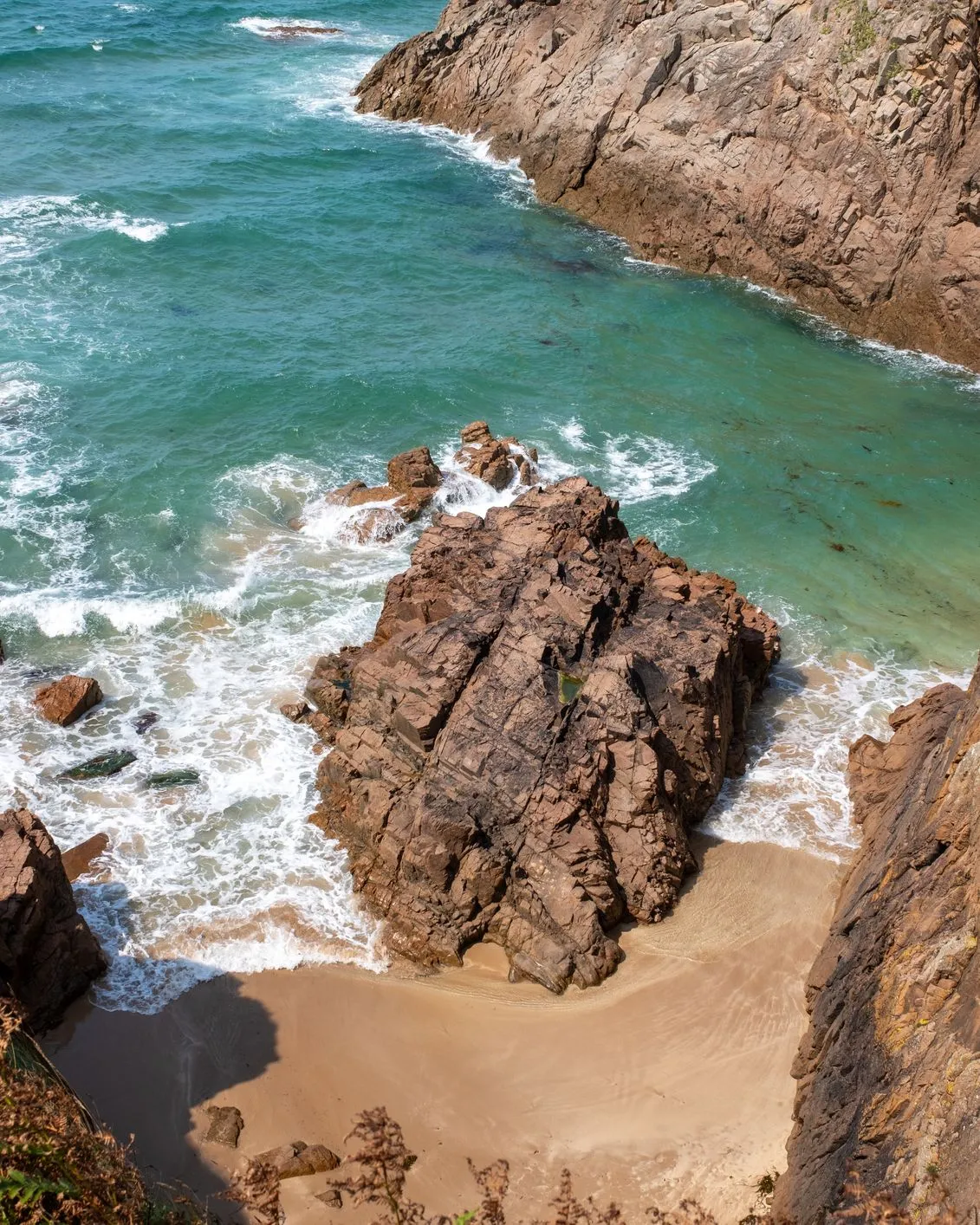Rocky coastal cove with turquoise sea, foaming waves and a small sandy beach below steep cliffs