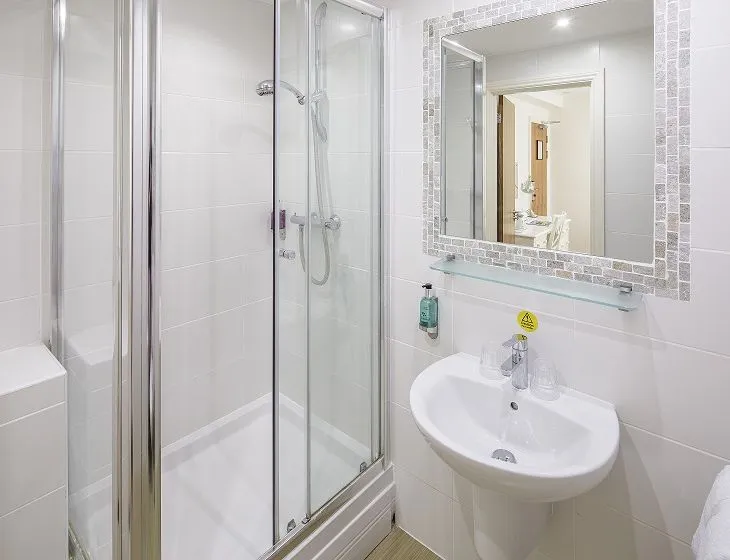 Modern white tiled bathroom with a glass shower enclosure, pedestal sink and illuminated wall mirror